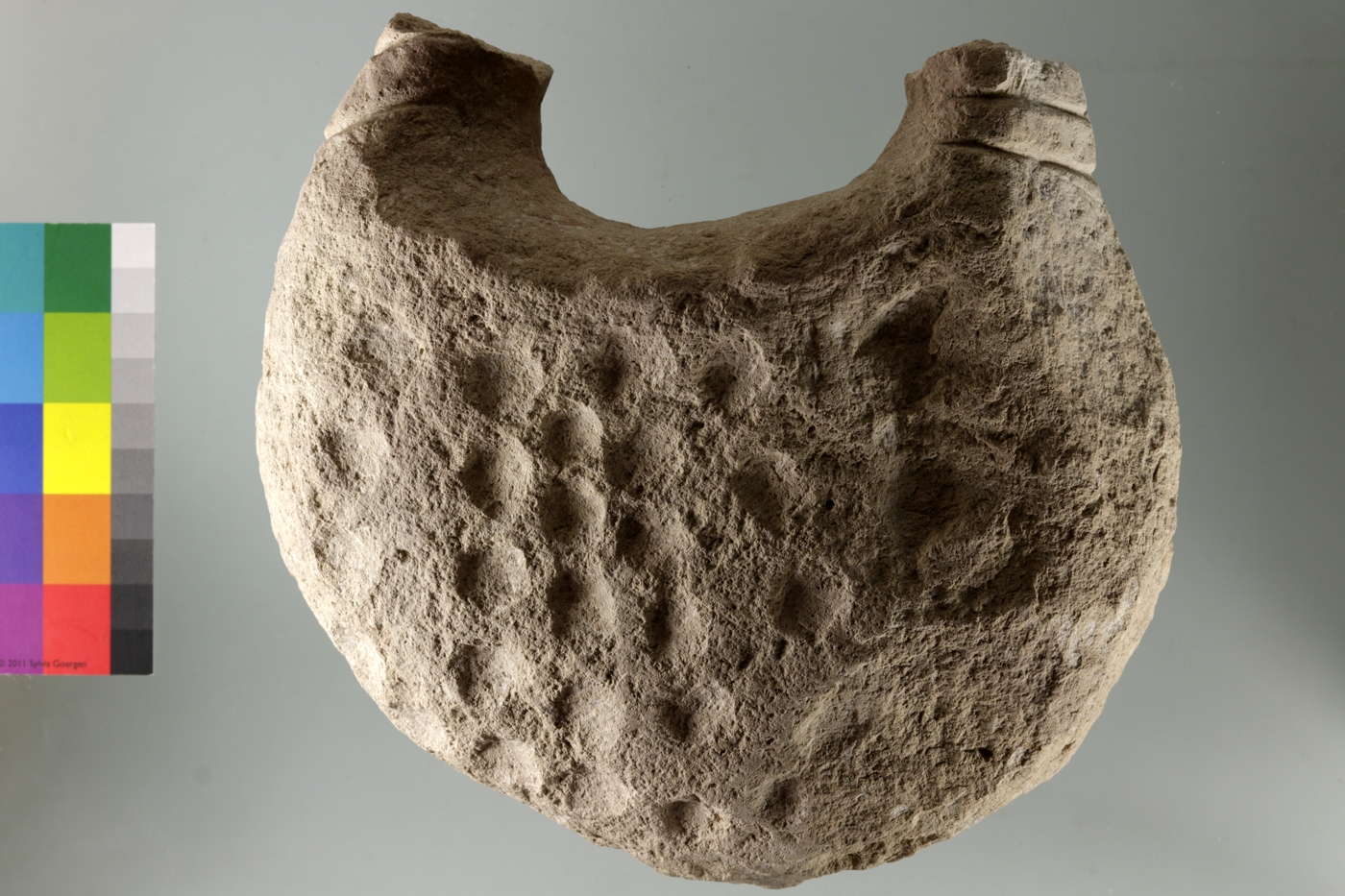 Ungewöhnliches „Handtaschen“-förmiges Steinobjekt. Die „Griffe“ sind an beiden Enden abgebrochen, der Körper des Objekts mit runden Vertiefungen bedeckt.